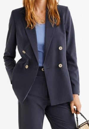 Vrouw die een donkerblauw double-breasted colbert draagt met beige knopen, donkerblauwe broek, lichtblauw shirt en een kleine gevlochten handtas vasthoudt.