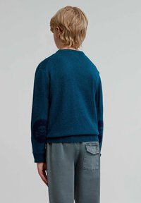 Suéter azul verdoso hecho de un tejido suave con cuello redondo y un diseño de calavera en el codo izquierdo, combinado con pantalones grises que tienen un bolsillo trasero.