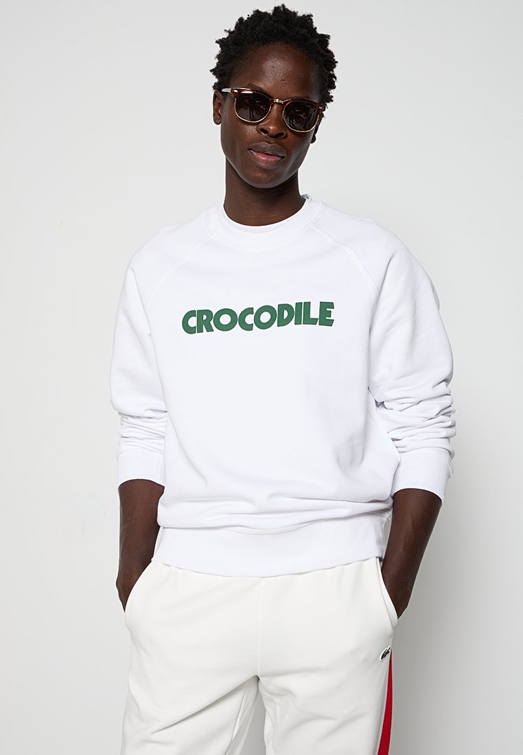 Weißer Hoodie mit grüner "CROCODILE"-Aufschrift. Verfügt über gerippte Bündchen und Saum, runden Halsausschnitt und eine lockere Passform. Getragen mit weißen Hosen.