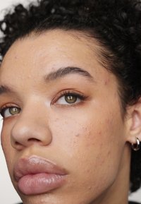 Gros plan sur un visage avec des cheveux bouclés brun clair, des yeux vert olive, et une texture de peau lisse et légèrement humide avec des imperfections visibles.