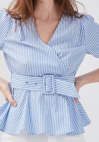 Kobieta ubrana w błękitną i białą bluzkę w poziome paski o kroju kopertowym z bufiastymi rękawami oraz dopasowanym szerokim paskiem z prostokątną klamrą.