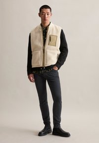 Man in zwarte jeans, zwarte laarzen, een zwart longsleeve shirt, een camouflage knoopshirt en een beige fleece vest met ritszakken.