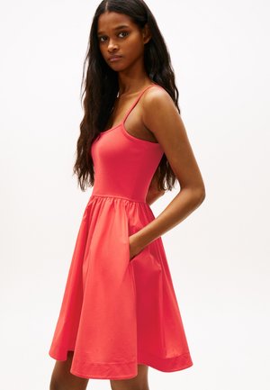 Kvinde med langt mørkt hår iført en ærmeløs koralrosa kjole med lommer, stående mod en ensfarvet hvid baggrund med den ene hånd i lommen.