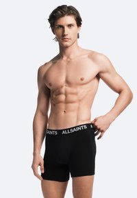 Schwarze Boxershorts mit einem breiten schwarzen Gummibund, der mit weißem "ALLSAINTS"-Branding versehen ist. Das Model hat einen muskulösen Körperbau und steht vor einem hellen Hintergrund.