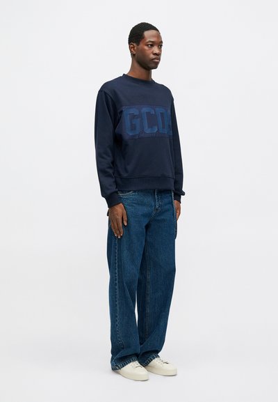 Marinblå sweatshirtdesign med stort, kontrasterande GCDS-logotyp. Löst sittande blå jeans. Modellen bär vita skor. Slät textur, avslappnad stil.