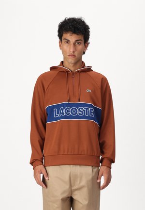Brun hettegenser med glidelås-krafter, med en blå stripe og hvit "LACOSTE" tekst over brystet. Laget av myk, teksturert stoff.