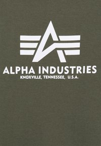 Logo di Alpha Industries con una "A" stilizzata e tre linee orizzontali, sopra il testo "Alpha Industries Knoxville, Tennessee, U.S.A." su tessuto verde.