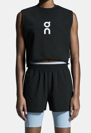 Person wearing sleeveless black cropped top with white logo and black shorts layered over light blue biker shorts against plain background.