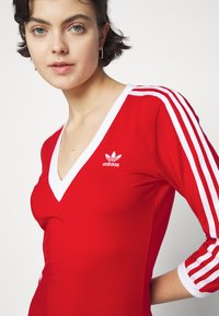 Rdeča športna majica z V-izrezom, belimi detajli in tremi belimi �črtami na vsaki rokavi. Ima vezen logotip Adidas na prsih.