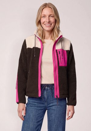 Femme portant une veste polaire à blocs de couleurs avec une fermeture éclair rose et une poche poitrine, chemise beige et jean bleu sur un fond neutre.