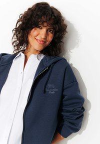 Veste à capuche zippée bleu marine avec une texture douce et un détail brodé "1975 Collection Audio" sur la poitrine ; associée à une chemise blanche à col.