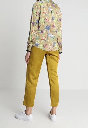 Blusa a fiori beige con disegni colorati di palme e fiori, abbinata a pantaloni a sigaretta giallo senape e sneaker bianche.