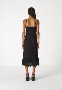 Vestido midaxi preto com corpete franzido, alças finas ajustáveis e uma bainha ondulada com babados, apresentando padrões recortados em ilhós.