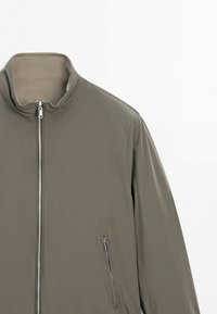 Light brown zip-up jacket with a high collar and zippered side pocket on the right, shown against a white background.