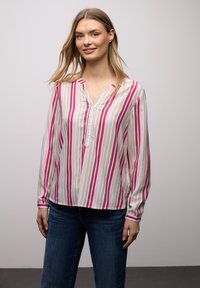 Femme souriante, portant un chemisier à manches longues avec des rayures verticales roses, blanches, beige et bleues, et un jean bleu foncé, devant un fond uni.