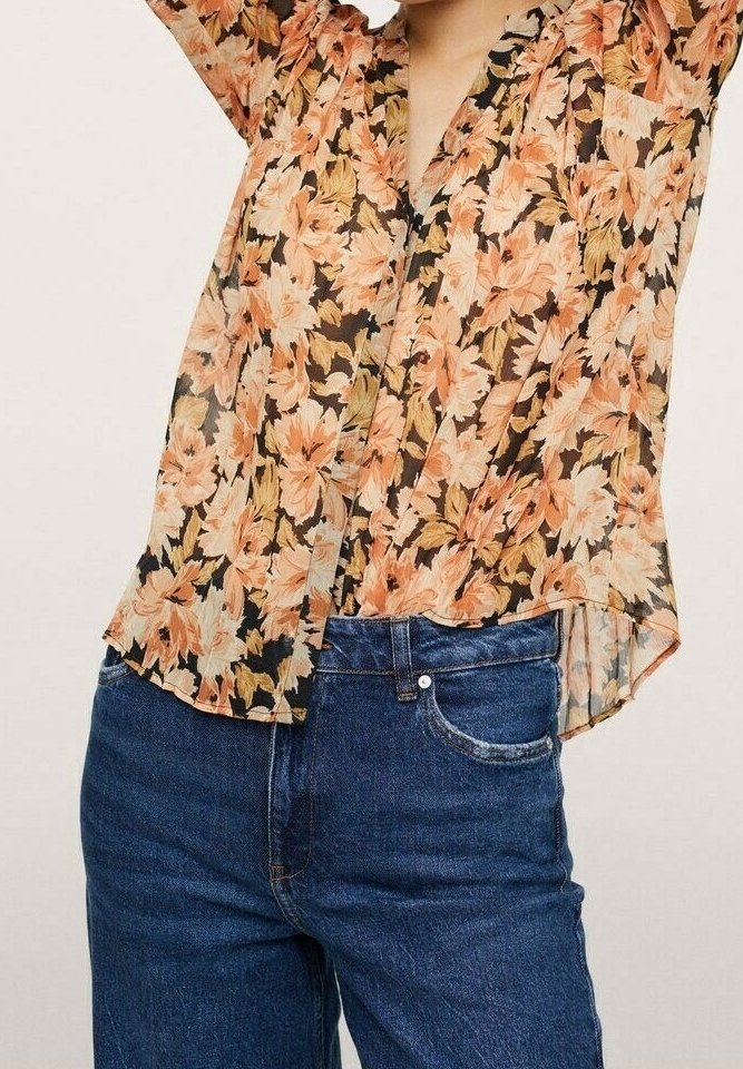 Blouse transparente à motif floral sur fond noir, ornée de fleurs orange et jaunes, à boutons et coupe ample, associée à un jean en denim bleu.