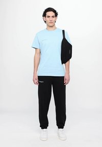 Light blue short-sleeve t-shirt with printed detail, worn with black sweatpants and a black crossbody bag. Model stands against a plain background.