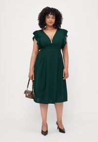 Robe verte, à mi-mollet, avec un décolleté en V profond et des manches bouffantes. Portée avec des escarpins noirs et un petit sac à main en imprimé animal.