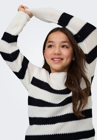Fille aux longs cheveux noirs souriante, portant un pull en tricot rayé noir et blanc, levant les deux bras au-dessus de la tête.