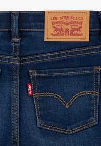 Los vaqueros de mezclilla azul oscuro presentan una etiqueta de cuero marrón en la cinturilla, una pestaña roja de Levi's en el bolsillo y costuras doradas que delinean la forma del bolsillo.