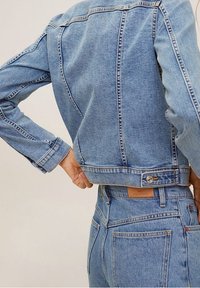 Veste en denim bleu clair avec une coupe raccourcie, des coutures visibles et des détails de boutons. Associée à un jean en denim taille haute, avec des poches arrière.