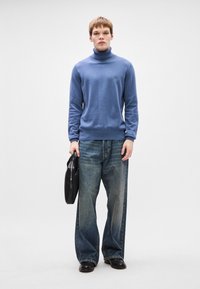 Pullover azul com gola alta, com punhos e bainha canelados, combinado com jeans azuis desbotados de corte largo e uma bolsa preta. Design simples e clean.