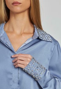 Camisa de satén azul claro con adornos de brillantes en el cuello y los puños. Textura suave con acentos metálicos para la decoración.
