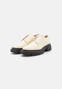 Scarpe in pelle bianca con una finitura liscia, caratterizzate da suole nere seghettate, punte rotonde e un design classico con lacci.