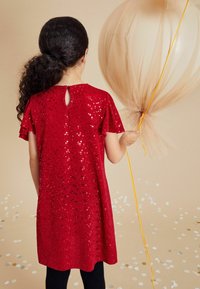 Robe rouge à sequins avec des manches courtes et flottantes, dos en forme de clé, associée à un ballon. Des leggings noirs complètent l'ensemble.