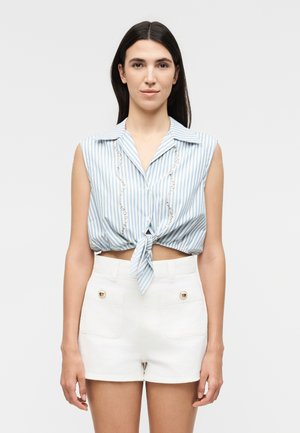 Γυναίκα με ίσια σκούρα μαλλιά φοράει αμάνικο μπλε και λευκό ριγέ πουκάμισο δετό μπροστά και λευκά σορτς ψηλόμεσα με τσέπες με κουμπιά.