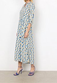 Abito maxi con uno sfondo beige chiaro caratterizzato da un motivo a foglie blu e nere. Maniche lunghe, scollatura con bottoni e forma fluente.