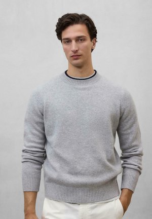 Suéter de punto gris claro con puños y dobladillo acanalados. Cuello redondo, textura suave. Llevado sobre una camisa oscura, exhibido contra un fondo neutro.