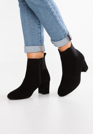 Person iført opsmøgede lyseblå jeans og sorte ruskind støvler med blokhæle, stående på et hvidt gulv.