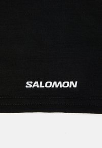 Svart t-skjorte i stoff med glatt tekstur, med en hvit "SALOMON"-logo trykket nær sømmen og rene sømdetaljer.