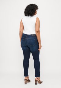 Dark blue skinny jeans with a mid-rise waist, featuring subtle whiskering, back pockets, and a frayed hem, paired with a white sleeveless top.