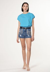 Blå kortærmet t-shirt parret med slidte denimshorts med kædeaccent og et sort bælte med en metallisk spænde. Hvide sandaler med stropper.