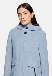 Helleblaue Wollmischungskapuze mit einer strukturierten Kapuze, zwei vorderen Taschen, schwarzen Knopfverschlüssen und einem kontrastierenden dunkelblauen Innenfutter.