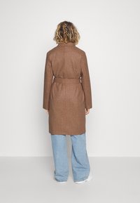 Brun, bælteholdt uldfrakke med krave og lange ærmer, parret med lyseblå brede bukser og hvide sneakers. Bagview vist.