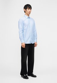 Νέος άνδρας όρθιος, φορέοντας ανοιχτό γαλάζιο πουκάμισο με κουμπιά, μαύρο παντελόνι φαρδιάς γραμμής και μαύρα επίσημα παπούτσια, σε απλό λευκό φόντο.