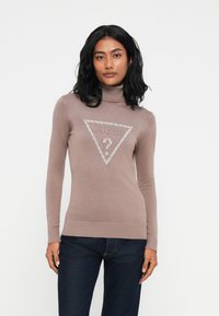 Donna con capelli neri che indossa un maglione a collo alto color malva con un logo GUESS lucido e jeans blu scuro, in piedi davanti a uno sfondo semplice.