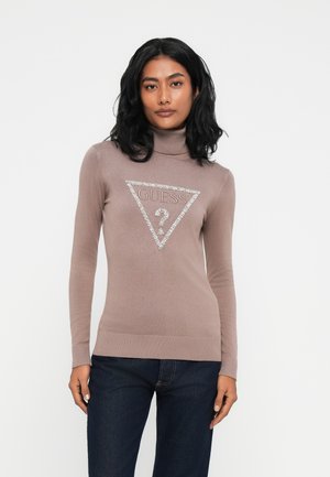 Donna con capelli neri che indossa un maglione a collo alto color malva con un logo GUESS lucido e jeans blu scuro, in piedi davanti a uno sfondo semplice.