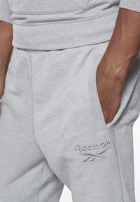 Nahaufnahme einer Hand, die sich in der rechten Tasche einer hellgrauen Jogginghose befindet, mit dem Reebok-Logo, das auf den Oberschenkel gestickt ist.