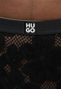 Μαύρο δαντελένιο ύφασμα με μοτίβο λουλουδιών και μαύρη μέση με ετικέτα λευκού λογότυπου "HUGO".