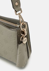 Coin d'un sac à main en cuir embossé gris avec une fermeture éclair dorée, une attache de sangle et un charm rond de la marque Guess fixé à l'anneau de la sangle.