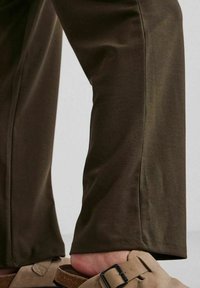 Oliveově zelené, slim-fit kalhoty vyrobené z měkké, elastické látky s hladkým povrchem. Na noze je vidět světle hnědé sandály s otevřenou špičkou.