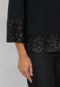 Schwarzes Oberteil mit einem Pailletten-Saum und Manschette, das einen glatten Stoff und glänzende Pailletten aufweist. Hand auf einem dunklen Denim-Stoff unten gezeigt.