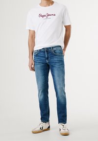 White t-shirt with red logo, blue denim jeans, and beige sneakers with navy and pink accents. Slim fit with a medium wash texture.