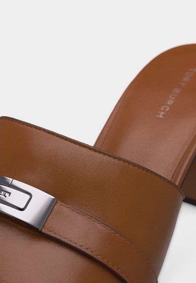 Καφέ δερμάτινο σανδάλι τύπου slide με ασημένια μεταλλική αγκράφα και χαμηλό τακούνι σε λευκό φόντο.