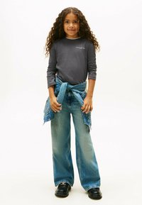 Mädchen mit langen lockigen Haaren, das ein dunkles langärmeliges Shirt, weite Jeans, eine um die Taille gebundene Jeansjacke und schwarze Schuhe trägt.