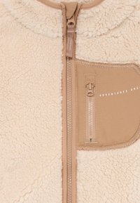 Casaco de fleece bege com uma textura suave, apresentando um fecho frontal e um bolso no peito com um zíper menor. Texto "AVENTURAS A CAMINHO" no bolso.